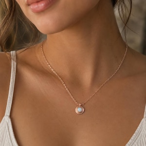 May include: A rose gold chain necklace featuring a small, circular pendant with a central iridescent white stone. The pendant has a textured, beaded border surrounding the stone, and the chain is a simple, delicate link style.