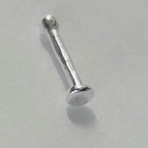 Pode incluir: Um piercing de nariz prateado com uma parte superior arredondada e uma base plana e circular. O piercing tem um design simples e elegante. O metal tem uma superfície polida e refletora.