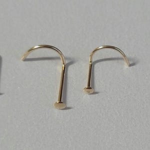 Puede incluir: Dos piercings de nariz dorados se muestran sobre un fondo gris claro. Los piercings tienen una parte superior curvada y un pequeño disco plano en la base. El metal tiene un acabado brillante.