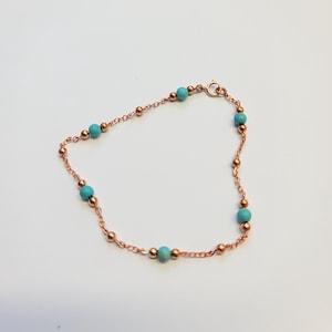 Puede incluir: Una delicada pulsera con una cadena de cobre con pequeñas cuentas turquesas y detalles en color cobre. La pulsera tiene un cierre de mosquetón. La joya se muestra sobre un fondo blanco.