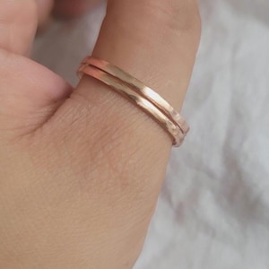 Puede incluir: Primer plano de dos anillos finos con textura martillada en un cálido color oro rosa. Los anillos están apilados en un dedo, mostrando su diseño simple y elegante. El fondo es una tela suave y neutra.