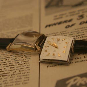 May include: Vintage gold-toned wristwatch with a square white face and gold numerals. The watch has a black leather band and is set against a backdrop of old newspaper text. The watch face reads "ELGIN".