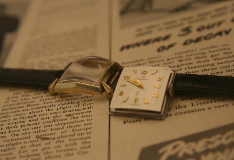 May include: Vintage gold-toned wristwatch with a square white face and gold numerals. The watch has a black leather band and is set against a backdrop of old newspaper text. The watch face reads "ELGIN".