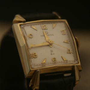 May include: A vintage gold-toned wristwatch with a square face and a black leather band. The watch face is white with gold-colored hands and hour markers. The watch reads "HEBROS" and "21".