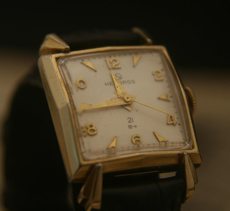 May include: A vintage gold-toned wristwatch with a square face and a black leather band. The watch face is white with gold-colored hands and hour markers. The watch reads "HEBROS" and "21".