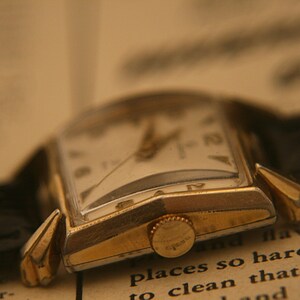 May include: Close-up of a vintage gold-toned wristwatch with a rectangular face and a black leather band. The watch has a white dial with black numerals and hands. The watch is resting on an open book with text visible.
