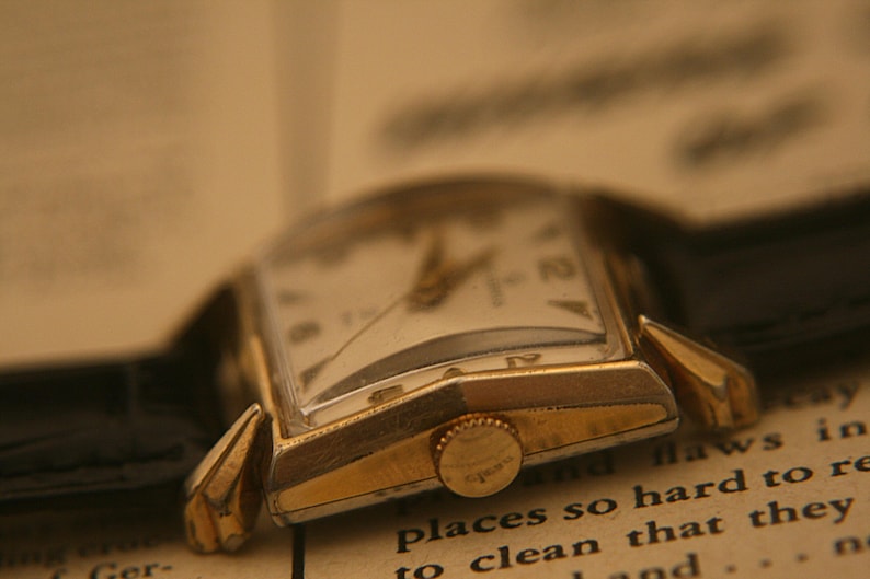 May include: Close-up of a vintage gold-toned wristwatch with a rectangular face and a black leather band. The watch has a white dial with black numerals and hands. The watch is resting on an open book with text visible.