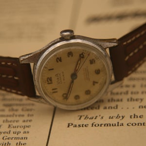 May include: A vintage wristwatch with a round silver-tone metal case, a cream-coloured dial featuring black Arabic numerals, and thin hands. The watch includes a brown leather strap with white stitching and the text Vira Geneve 17 Rubis on the dial.