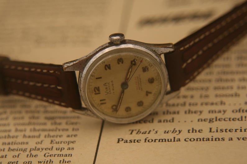 May include: A vintage wristwatch with a round silver-tone metal case, a cream-coloured dial featuring black Arabic numerals, and thin hands. The watch includes a brown leather strap with white stitching and the text Vira Geneve 17 Rubis on the dial.