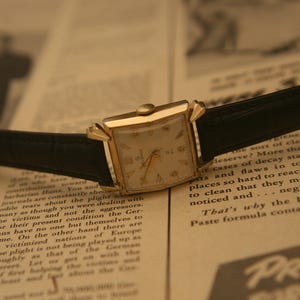 May include: A vintage gold wristwatch with a rectangular face and black leather band rests on a newspaper. The watch has a cream-colored dial with gold hands and hour markers. The newspaper's text is visible.