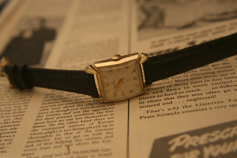 May include: A vintage gold wristwatch with a rectangular face and black leather band rests on a newspaper. The watch has a cream-colored dial with gold hands and hour markers. The newspaper's text is visible.