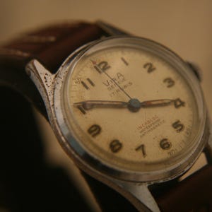 May include: A vintage Vira Geneve wristwatch with a round silver-tone case, a cream-coloured dial featuring black Arabic numerals, and a brown leather strap. The dial displays the text Vira Geneve, 17 Rubis, Incabloc, Waterproof, and Antimagnetic.
