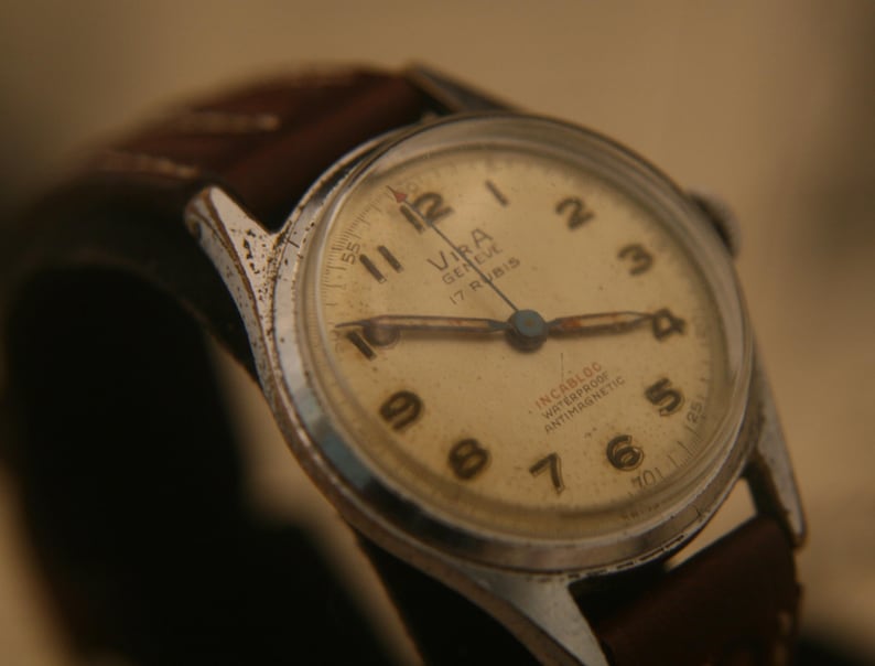 May include: A vintage Vira Geneve wristwatch with a round silver-tone case, a cream-coloured dial featuring black Arabic numerals, and a brown leather strap. The dial displays the text Vira Geneve, 17 Rubis, Incabloc, Waterproof, and Antimagnetic.