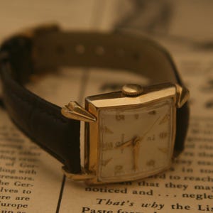 May include: A vintage gold-toned wristwatch with a rectangular face and a black leather band. The watch has gold hands and hour markers. The watch is resting on a printed paper surface.