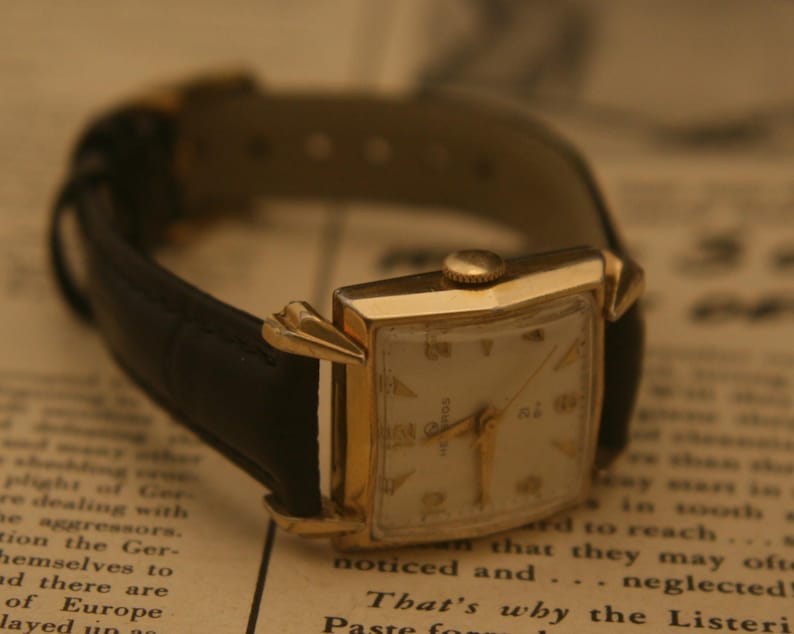 May include: A vintage gold-toned wristwatch with a rectangular face and a black leather band. The watch has gold hands and hour markers. The watch is resting on a printed paper surface.