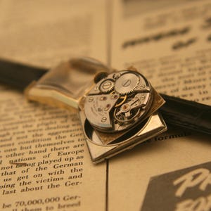 May include: Close-up of a vintage wristwatch with exposed gears and a black leather band. The watch has a gold-toned case and is set against a backdrop of aged paper with printed text. The watch mechanism is visible.