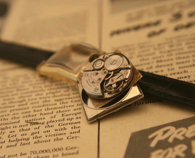 May include: Close-up of a vintage wristwatch with exposed gears and a black leather band. The watch has a gold-toned case and is set against a backdrop of aged paper with printed text. The watch mechanism is visible.