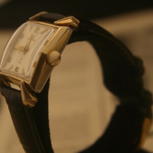 May include: A vintage gold-toned wristwatch with a rectangular face and a black leather band. The watch face is cream-colored with gold hands and numerals. The watch is angled, showcasing its design and craftsmanship.