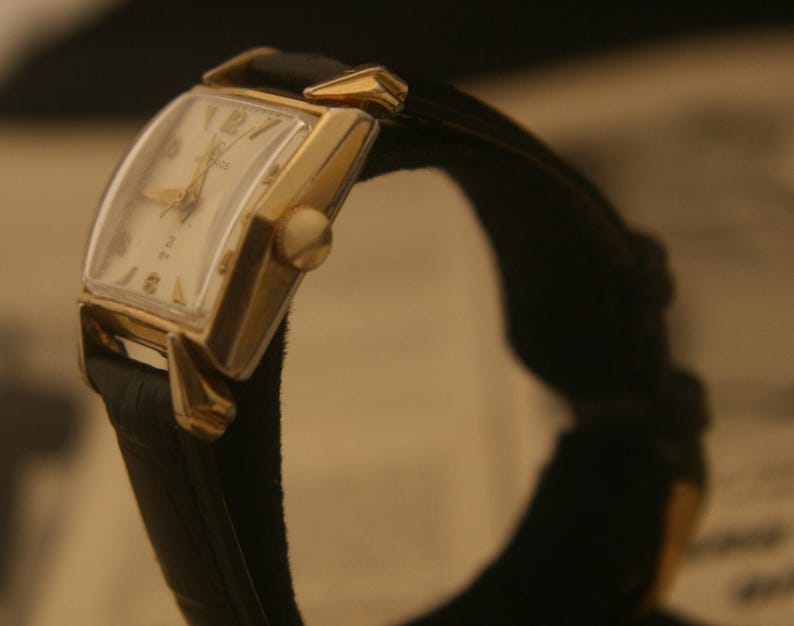 May include: A vintage gold-toned wristwatch with a rectangular face and a black leather band. The watch face is cream-colored with gold hands and numerals. The watch is angled, showcasing its design and craftsmanship.