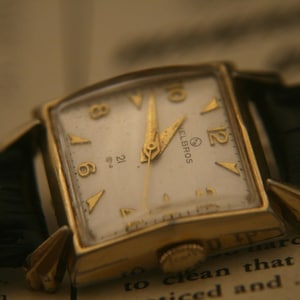 May include: A vintage gold-toned Helbros wristwatch with a square face and a white dial. The watch features gold hands and hour markers, and a black leather strap. The watch is set against a backdrop of aged paper.