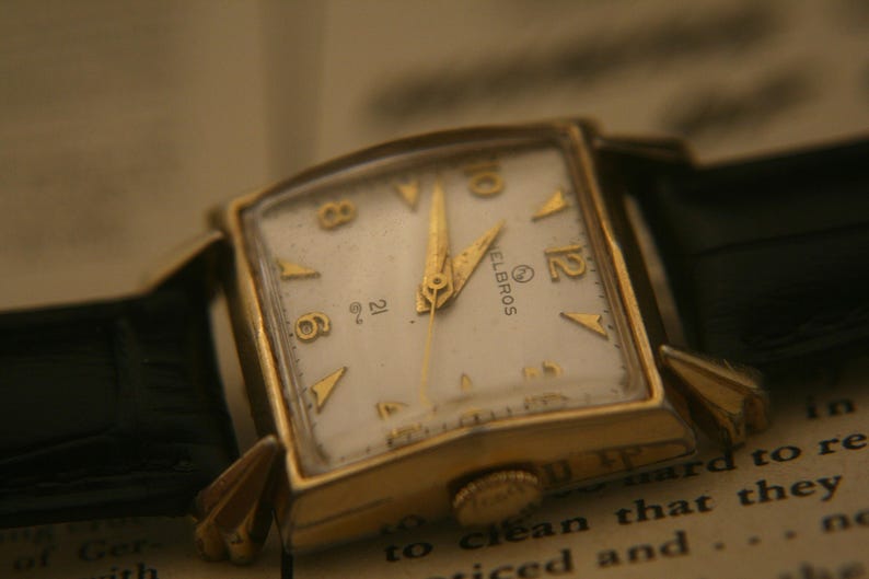 May include: A vintage gold-toned Helbros wristwatch with a square face and a white dial. The watch features gold hands and hour markers, and a black leather strap. The watch is set against a backdrop of aged paper.