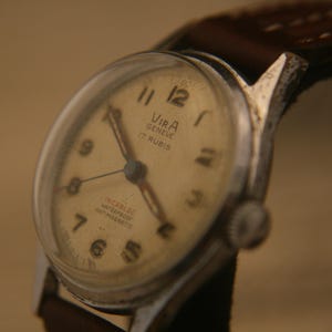 May include: A vintage wristwatch with a silver-toned metal case and a brown leather strap. The off-white dial features black Arabic numerals, silver hands, and the text VIRA GENEVE 17 RUBIS, INCABLOC, WATERPROOF, and ANTIMAGNETIC printed in black.