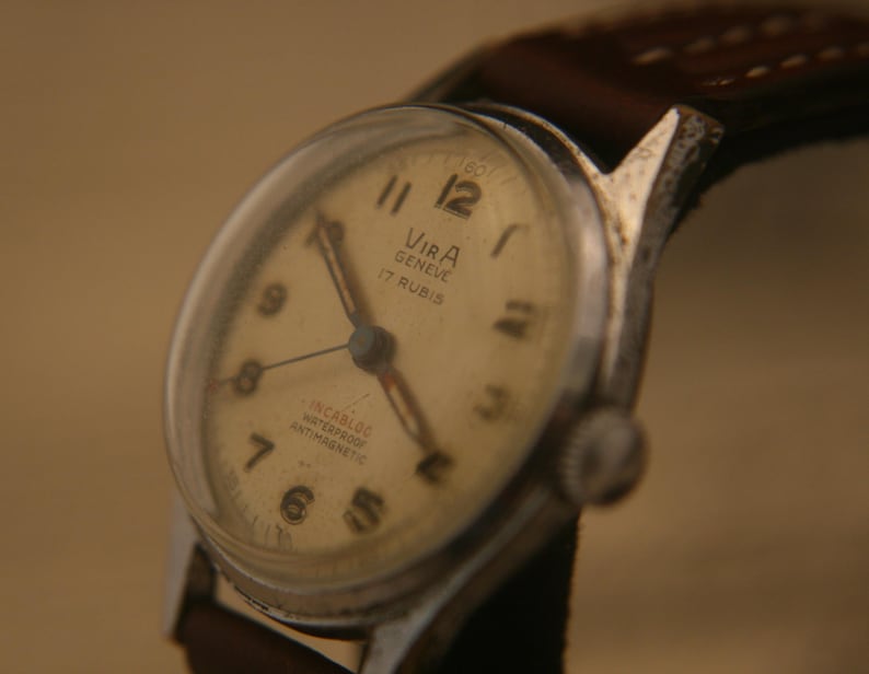 May include: A vintage wristwatch with a silver-toned metal case and a brown leather strap. The off-white dial features black Arabic numerals, silver hands, and the text VIRA GENEVE 17 RUBIS, INCABLOC, WATERPROOF, and ANTIMAGNETIC printed in black.