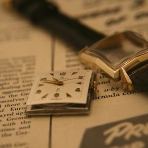 May include: Close-up of a vintage gold wristwatch with a black leather band, and a detached watch face. The watch face is square with gold hands and black numerals. The background is a newspaper.