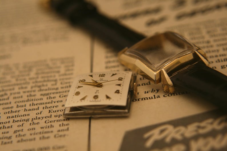 May include: Close-up of a vintage gold wristwatch with a black leather band, and a detached watch face. The watch face is square with gold hands and black numerals. The background is a newspaper.