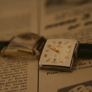 May include: Vintage wristwatch with a square, cream-colored face and gold-tone hands and numerals. The watch has a black leather band and is set against a backdrop of aged newspaper text.