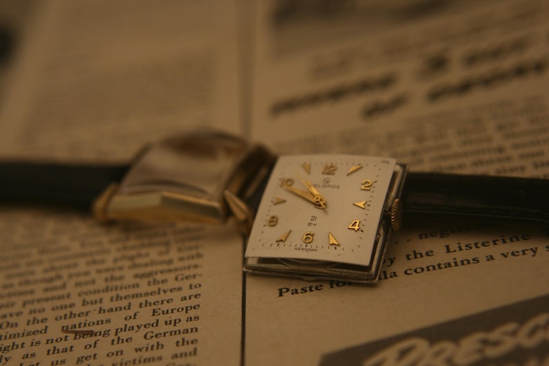 May include: Vintage wristwatch with a square, cream-colored face and gold-tone hands and numerals. The watch has a black leather band and is set against a backdrop of aged newspaper text.