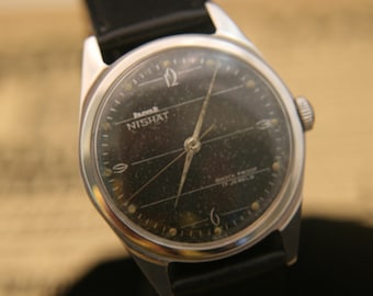 Könnte beinhalten: Eine silberfarbene Armbanduhr mit schwarzem Zifferblatt, auf dem HMT NISHAT, SHOCK PROOF und 17 JEWELS steht. Die Uhr hat ein schwarzes Lederarmband, silberne Zeiger und Ziffern an den Positionen 12, 6 und 9 auf dem Zifferblatt.