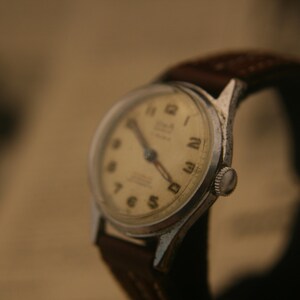May include: A vintage wristwatch with a silver-toned metal case and a cream-coloured dial featuring black Arabic numerals. The watch includes dark-coloured hands and is attached to a dark brown leather strap with visible stitching along the edges.