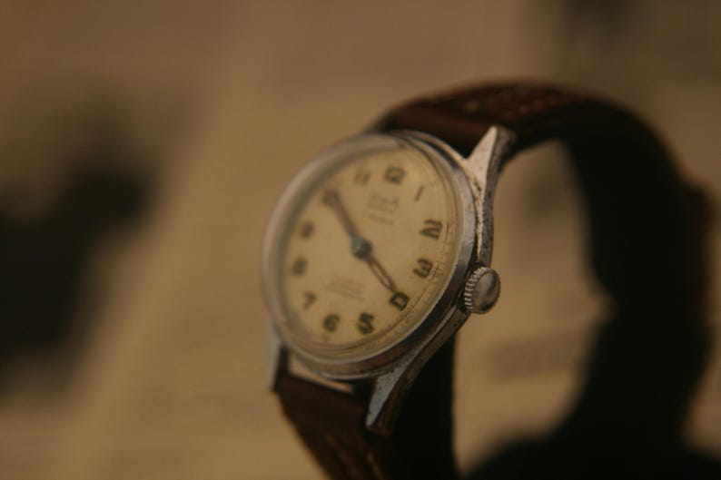 May include: A vintage wristwatch with a silver-toned metal case and a cream-coloured dial featuring black Arabic numerals. The watch includes dark-coloured hands and is attached to a dark brown leather strap with visible stitching along the edges.