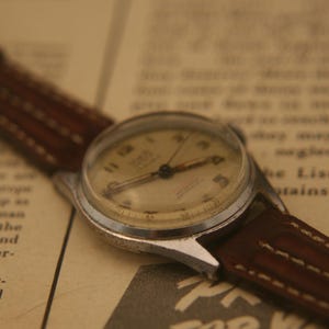 May include: A vintage wristwatch with a round silver-toned metal case and a cream-coloured dial featuring dark numerals. The watch is attached to a brown leather strap with visible stitching along the edges, showcasing a classic analogue design.