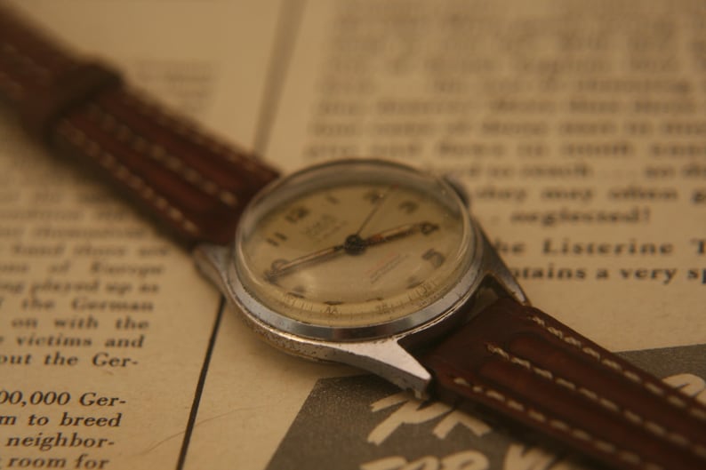 May include: A vintage wristwatch with a round silver-toned metal case and a cream-coloured dial featuring dark numerals. The watch is attached to a brown leather strap with visible stitching along the edges, showcasing a classic analogue design.
