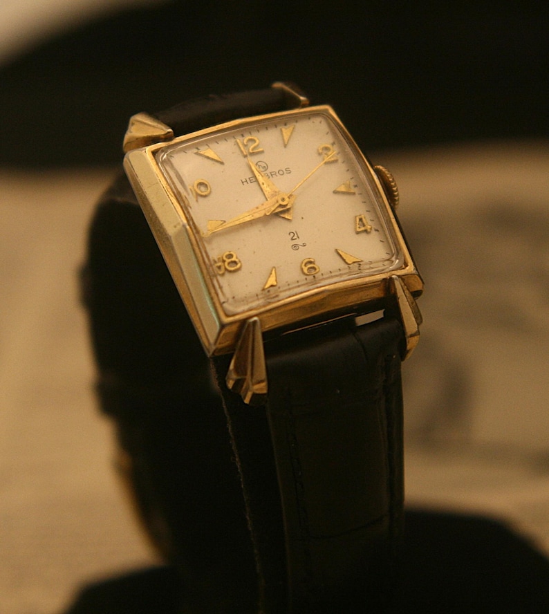 May include: A vintage gold-toned wristwatch with a rectangular face and a black leather band. The watch face is white with gold hands and numerals. The brand name "HEBROS" is visible on the watch face.