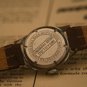 May include: A silver-toned wristwatch with a brown leather strap. The back of the watch case is stainless steel and features the text "STAINLESS STEEL BACK ANTIMAGNETIC WATERPROTECTED SHOCK PROTECTED INCABLOC" engraved in a circular pattern.
