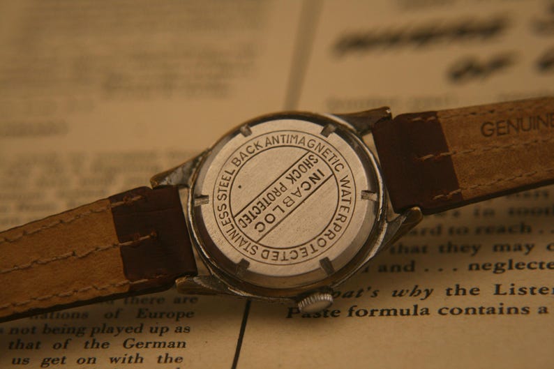 May include: A silver-toned wristwatch with a brown leather strap. The back of the watch case is stainless steel and features the text "STAINLESS STEEL BACK ANTIMAGNETIC WATERPROTECTED SHOCK PROTECTED INCABLOC" engraved in a circular pattern.