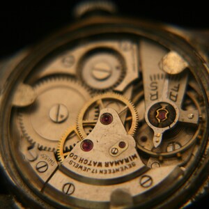May include: A mechanical watch movement with an exposed gear train and balance wheel. The metal plate is engraved with "SEVENTEEN 17 JEWELS MIRAMAR WATCH CO" and "SWISS". The mechanism features gold-toned gears, ruby bearings, and silver-toned bridges.