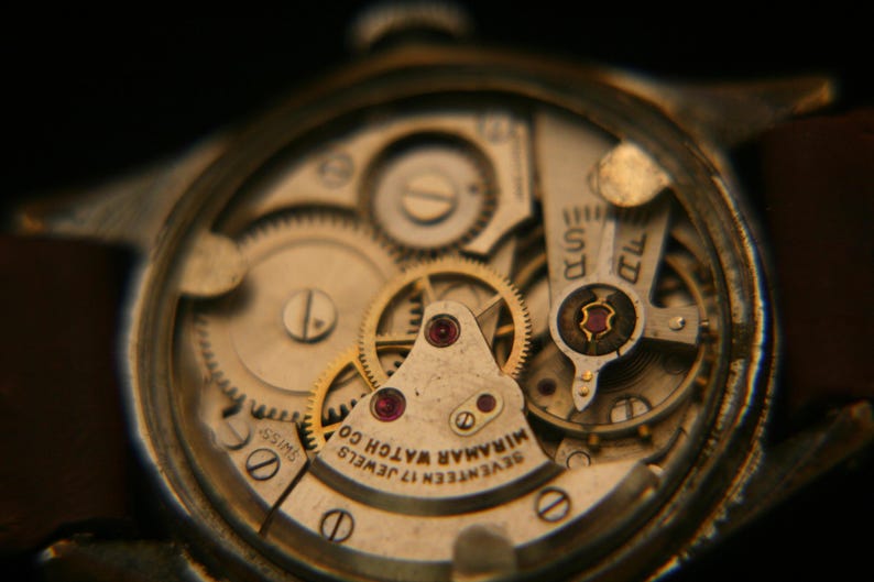 May include: A mechanical watch movement with an exposed gear train and balance wheel. The metal plate is engraved with "SEVENTEEN 17 JEWELS MIRAMAR WATCH CO" and "SWISS". The mechanism features gold-toned gears, ruby bearings, and silver-toned bridges.