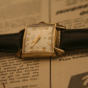 May include: Vintage gold-toned wristwatch with a rectangular face and a white dial. The watch features gold hands and hour markers, and a black leather band. The watch is resting on a printed paper background.