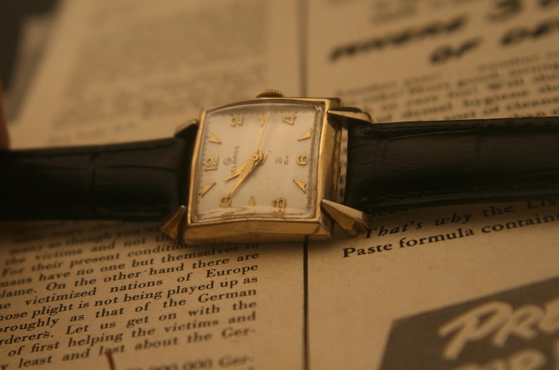 May include: Vintage gold-toned wristwatch with a rectangular face and a white dial. The watch features gold hands and hour markers, and a black leather band. The watch is resting on a printed paper background.