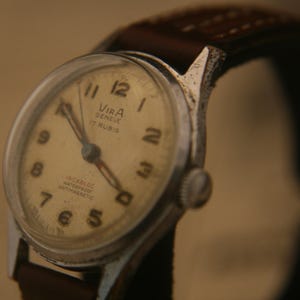 May include: A Vira Geneve wristwatch with a cream-coloured dial, black Arabic numerals, and silver-tone hands. The watch face features the text Vira Geneve 17 Rubis, Incabloc, Waterproof, and Antimagnetic. It has a brown leather strap with stitching.