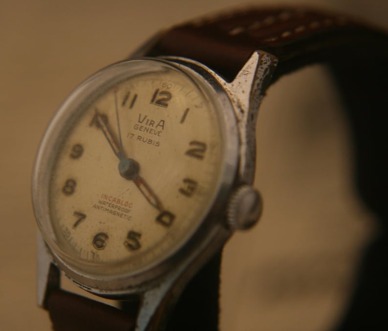 May include: A Vira Geneve wristwatch with a cream-coloured dial, black Arabic numerals, and silver-tone hands. The watch face features the text Vira Geneve 17 Rubis, Incabloc, Waterproof, and Antimagnetic. It has a brown leather strap with stitching.