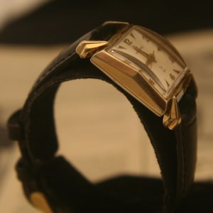 May include: A vintage wristwatch with a rectangular gold-toned case and a black leather band. The watch face is white with black numerals and hands. The watch is displayed at an angle, showcasing its classic design.