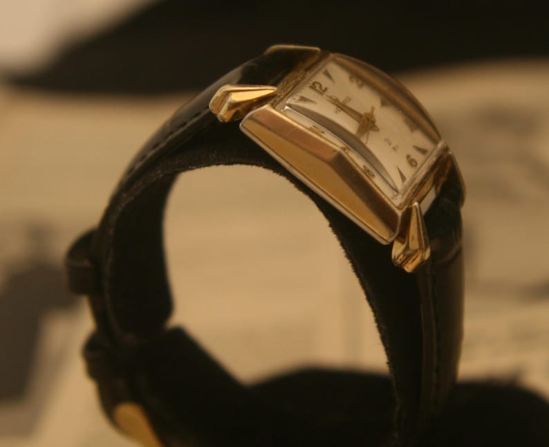 May include: A vintage wristwatch with a rectangular gold-toned case and a black leather band. The watch face is white with black numerals and hands. The watch is displayed at an angle, showcasing its classic design.