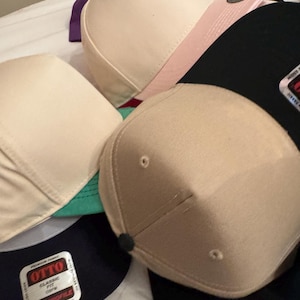 May include: A collection of baseball caps in various colors, including tan, black, pink, and purple. Some caps have a classic fit and a mid-profile design. The caps are made of fabric and have a structured shape.