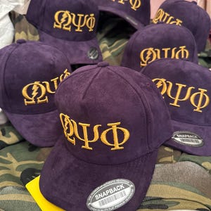May include: A collection of purple snapback hats with gold embroidered Greek letters. The hats have a flat brim and a snapback closure. The hats are labeled "SNAPBACK" and "THE PIONEER OF AUTHENTIC HEADWEAR HIGH QUALITY FLAT PREMIUM HEADWEAR".