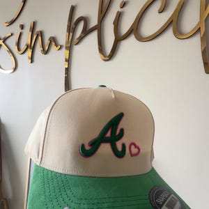 Puede incluir: Gorra de béisbol beige y verde con la letra "A" en verde con contorno rosa y un corazón rosa. La gorra tiene una visera verde y cierre snapback.